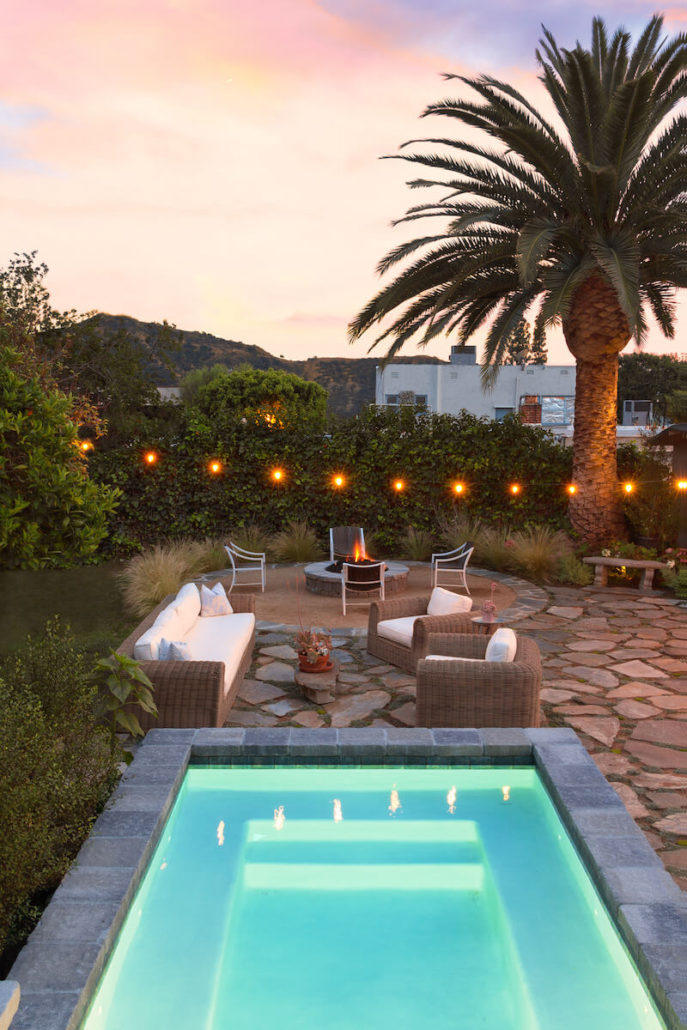 Twilight patio with a glowing turquoise pool, palm tree, lounge chairs, fire pit, string lights, and hills creating a serene retreat.