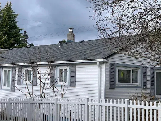 A single-story home displays white siding with gray shutters, a dark shingle roof, white fencing, ma 