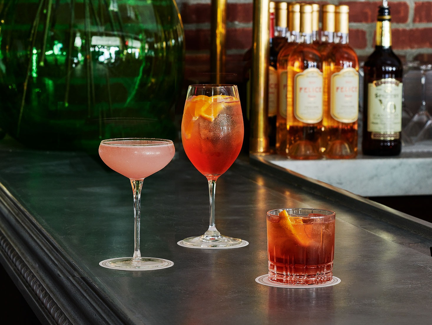 A pink coupe cocktail, an orange spritz, and a dark red Negroni are lined up on a dark bar counter. In the background, bottles of wine and a large green glass jug are visible.