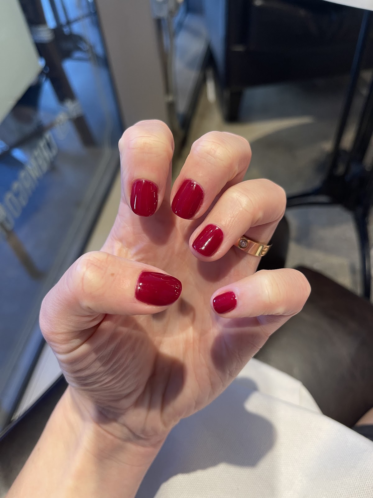 A close-up shot of a woman's hand with neatly manicured nails painted a deep red color. She is wearing a gold ring with a small diamond on her ring finger. The background is blurred but appears to be an indoor setting, possibly a salon or a well-lit room.