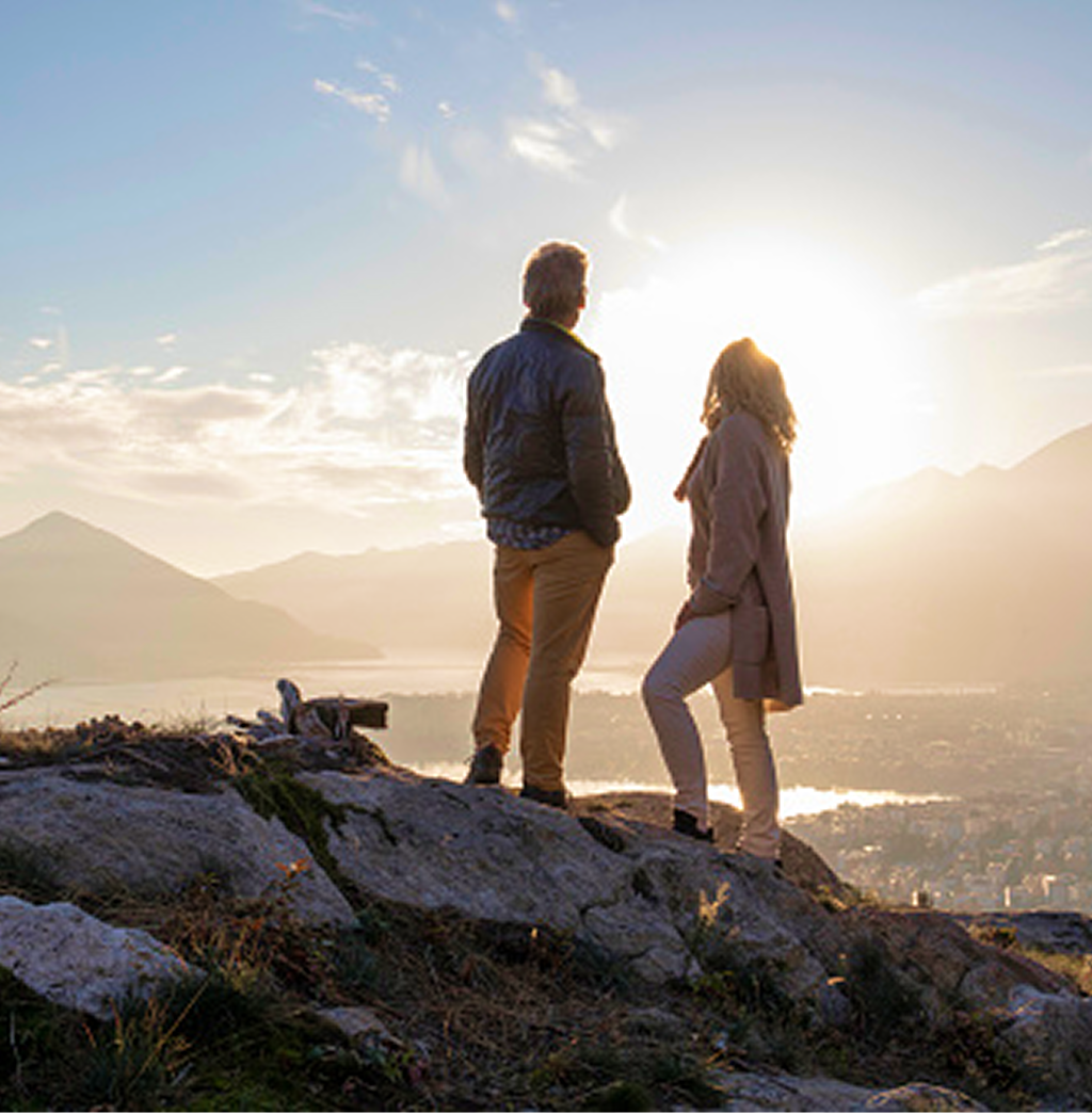 A man and woman standing on a rocky hilltop and looking out at the view and sunrise.