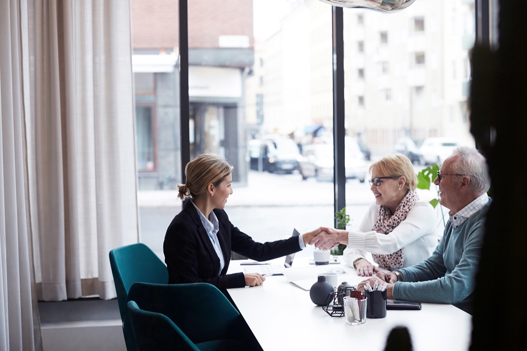 A woman in a business suit shakes hands with an older woman at a table in a modern office. An older man watches, and large windows show a city street.
