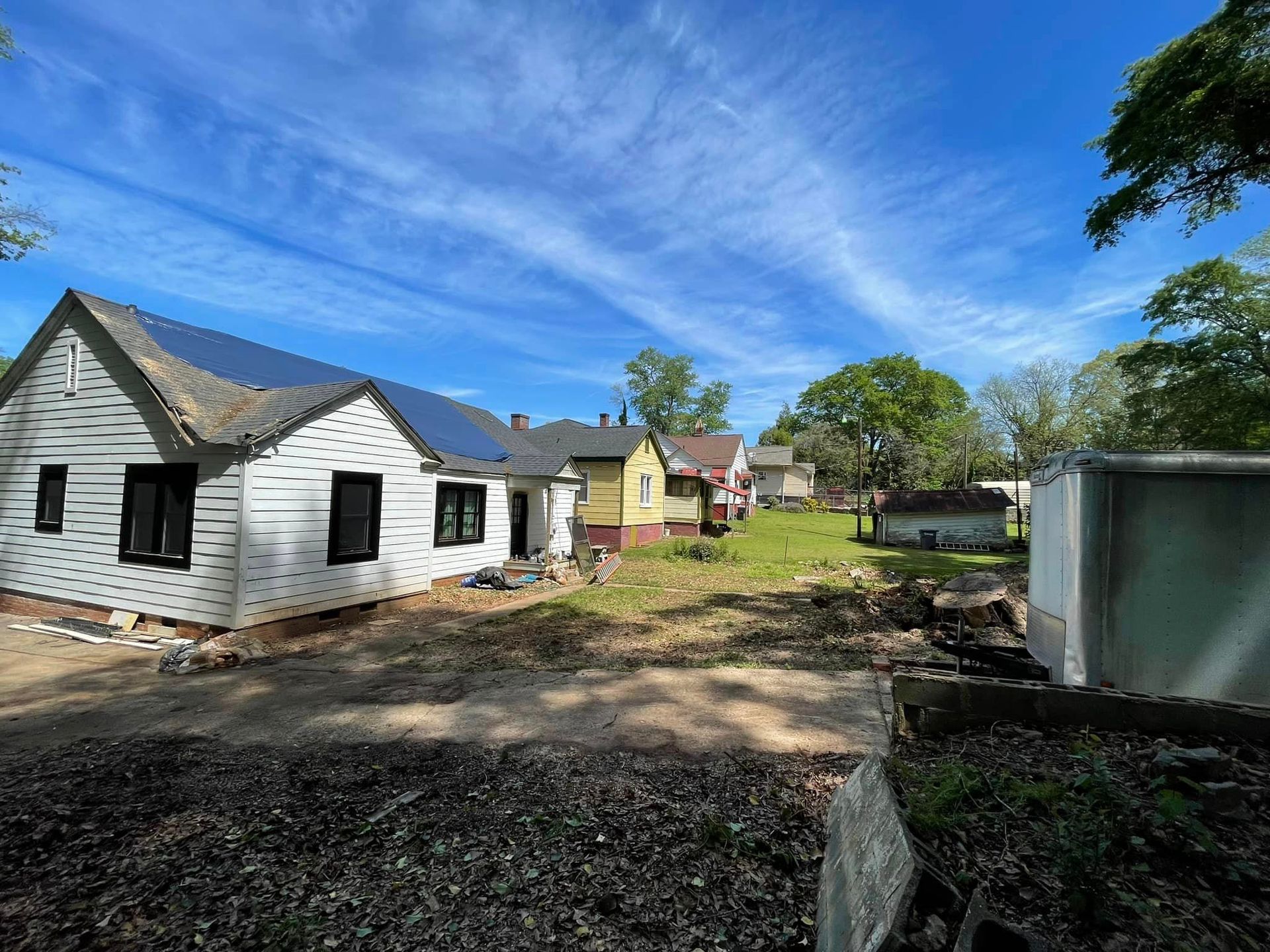 Residential neighborhood with multiple small houses, one featuring solar panels on the roof, open yard space, trees, and a trailer nearby, under a bright blue sky with wispy clouds.