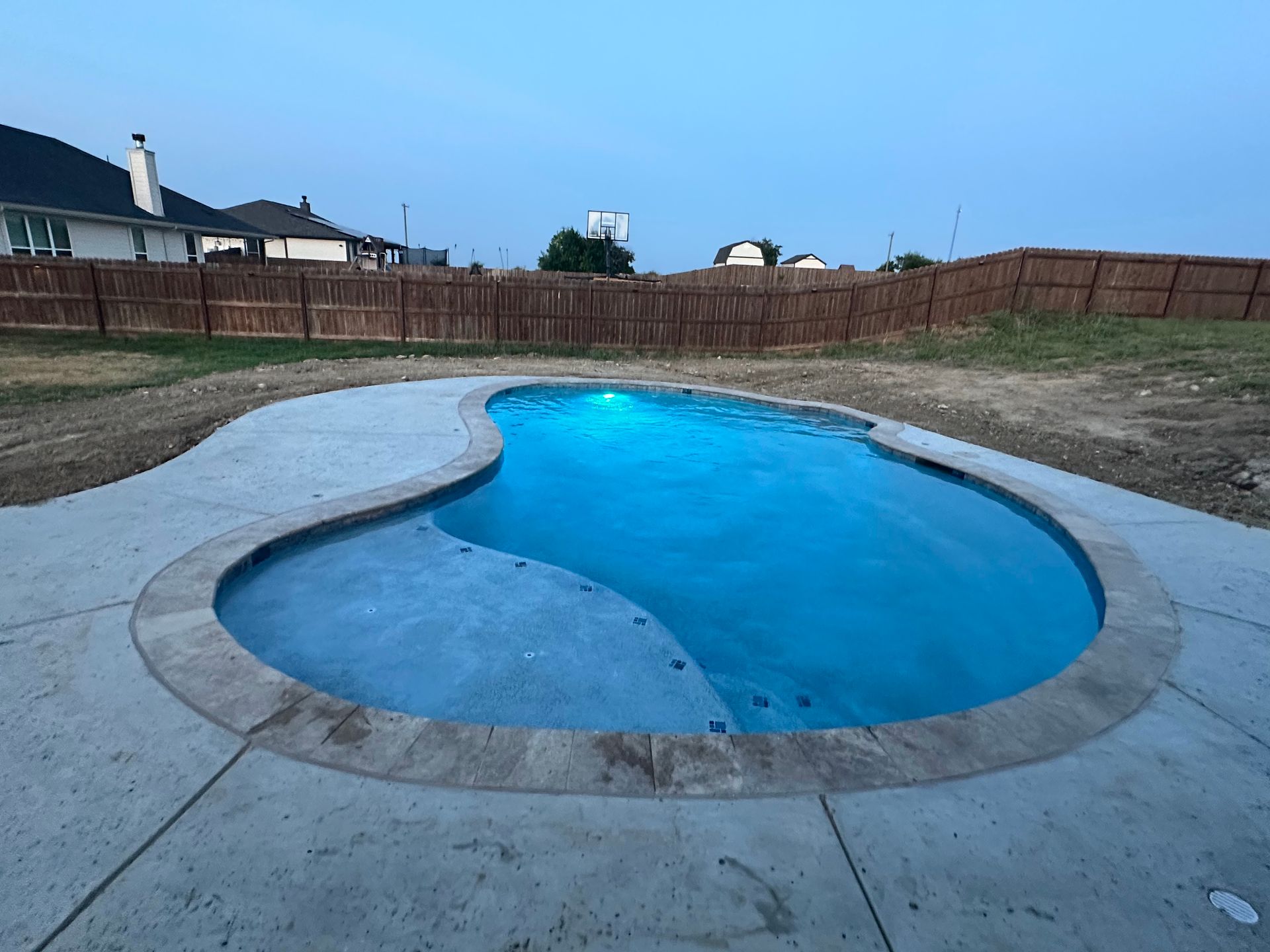 This finished freeform pool glows with internal blue lighting during twilight. The design features a large tanning ledge and smooth concrete decking, all contained within a private fenced-in backyard.