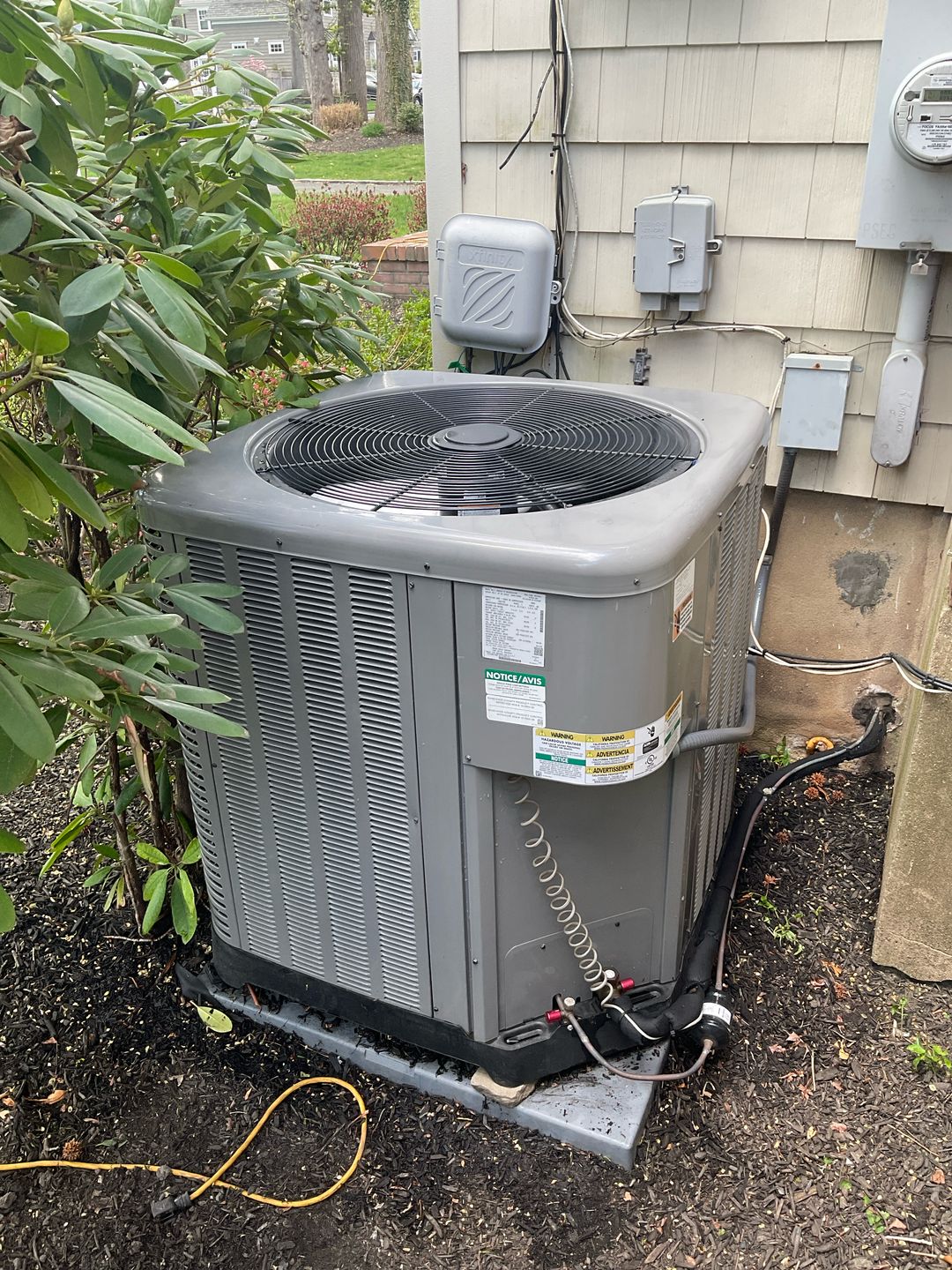 An outdoor air conditioning unit sits next to a house, partially obscured by a leafy green bush. The unit is gray and rectangular with a large fan on top. Various wires and electrical boxes are attached to the side of the house near the unit. The ground around the unit is covered in dark mulch, and a yellow electrical cord is visible on the ground.