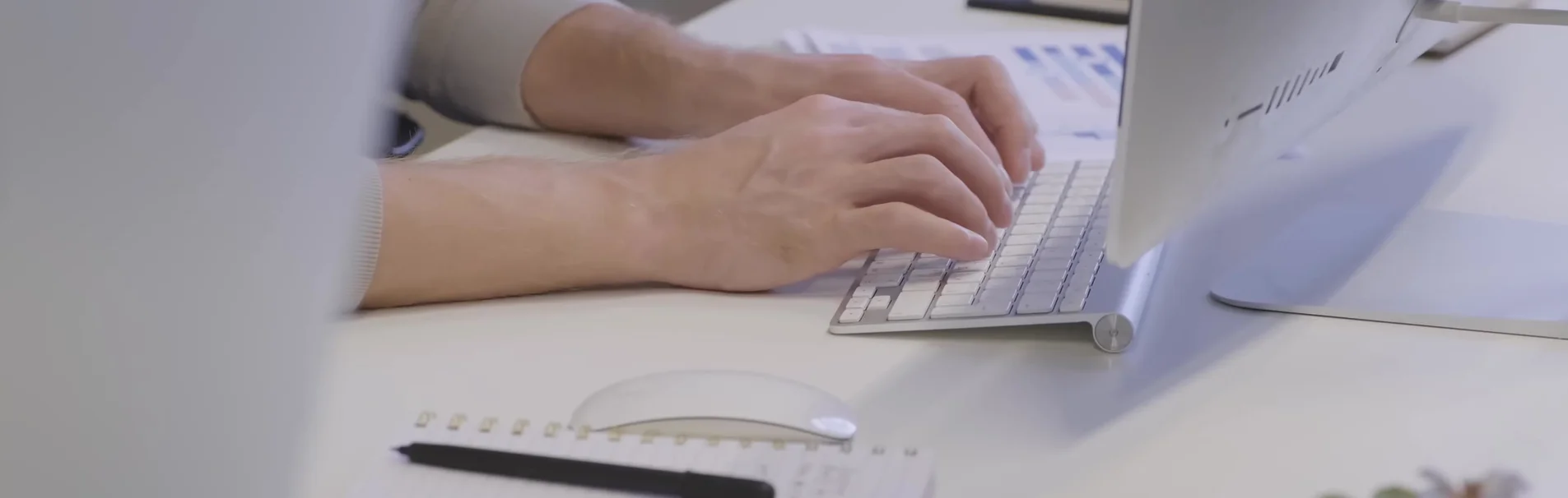 A person's hands typing on a computer keyboard in an office setting. A white mouse and a notebook with a pen are visible on the desk.