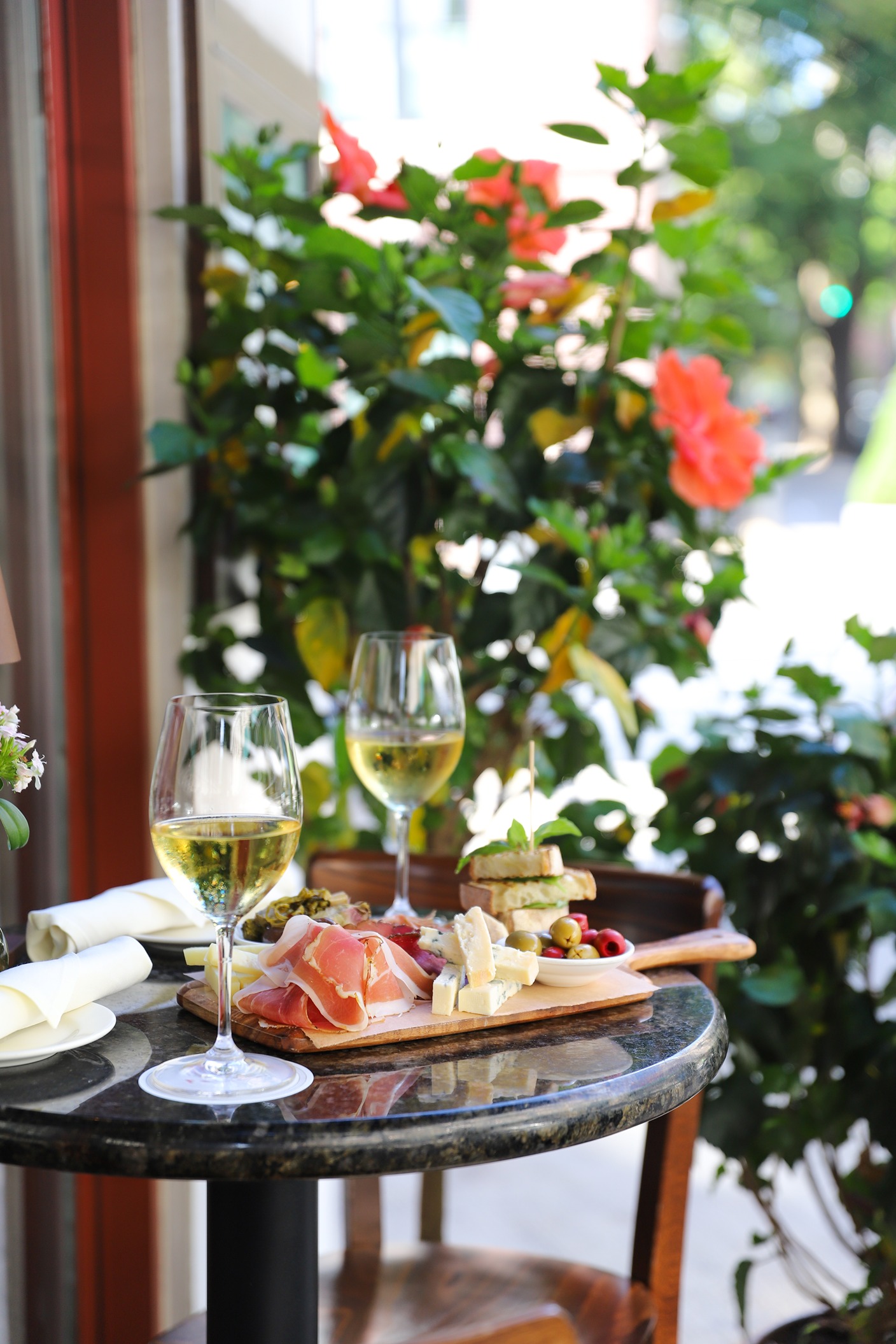 An Al fresco dining experience featuring a wooden board of prosciutto, cheeses, and olives next to blooming hibiscus flowers.