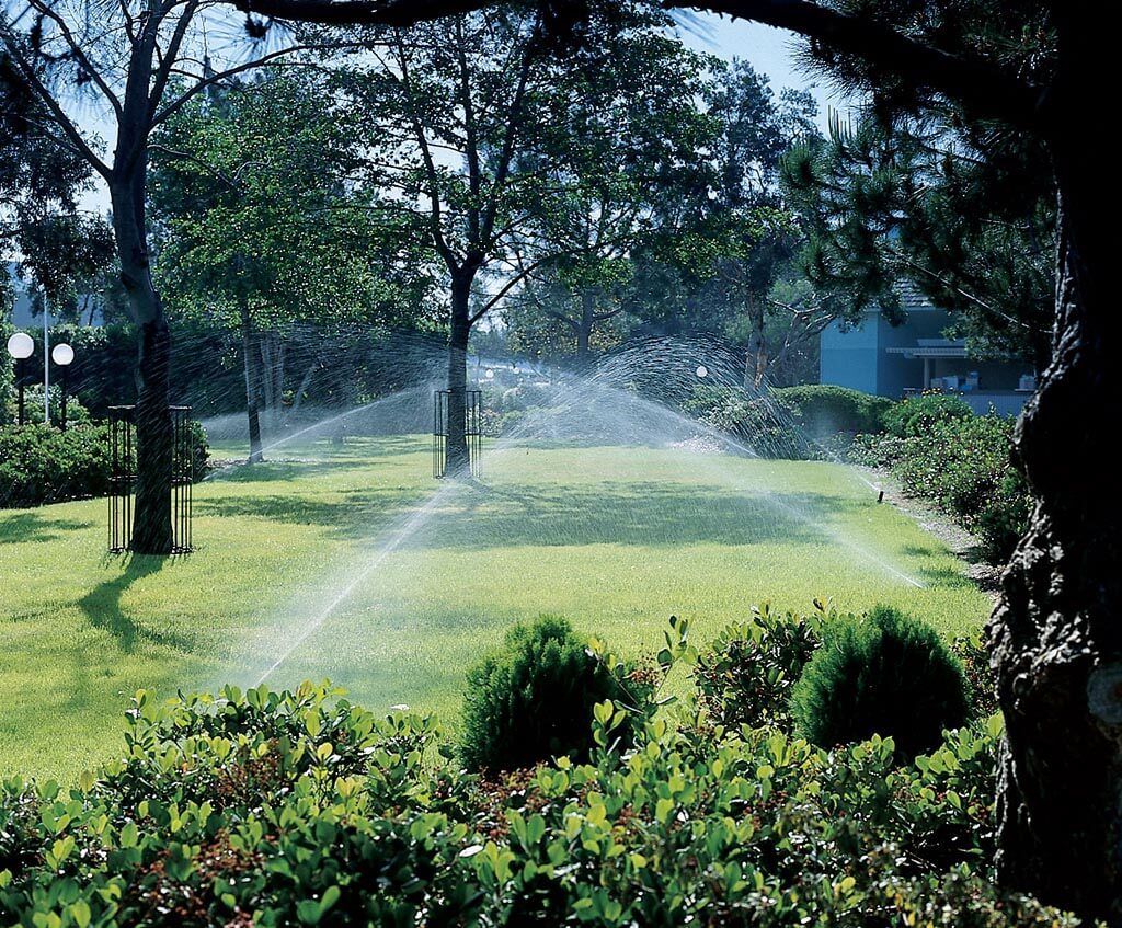 Multiple active sprinkler zones misting water across a large, beautifully landscaped lawn surrounded by mature trees.