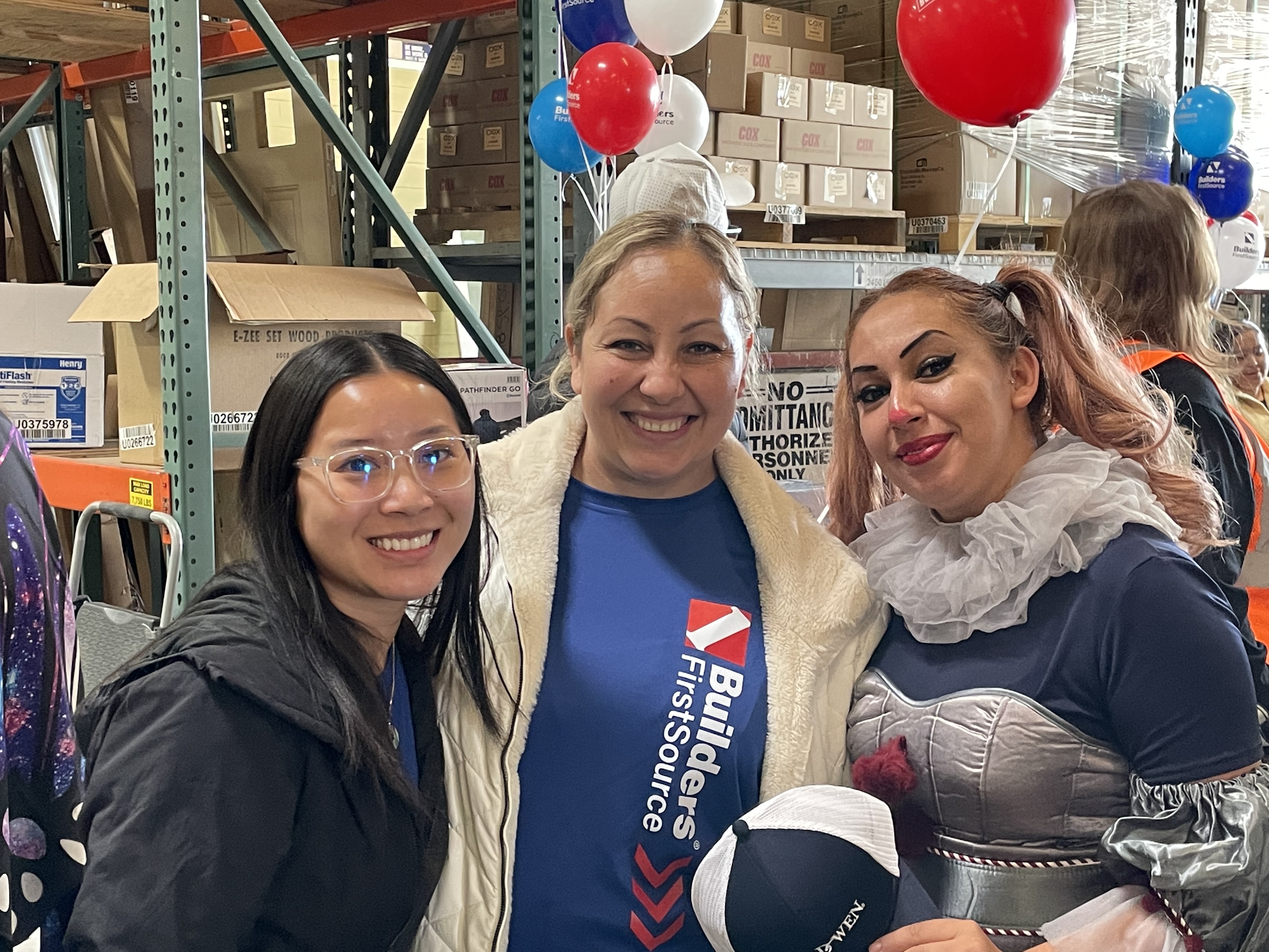 Three women are standing together, smiling at the camera. One woman is wearing a "Builders FirstSource" t-shirt, another is dressed in a costume with makeup, and the third is wearing glasses and a black jacket. Balloons and warehouse shelves are visible in the background