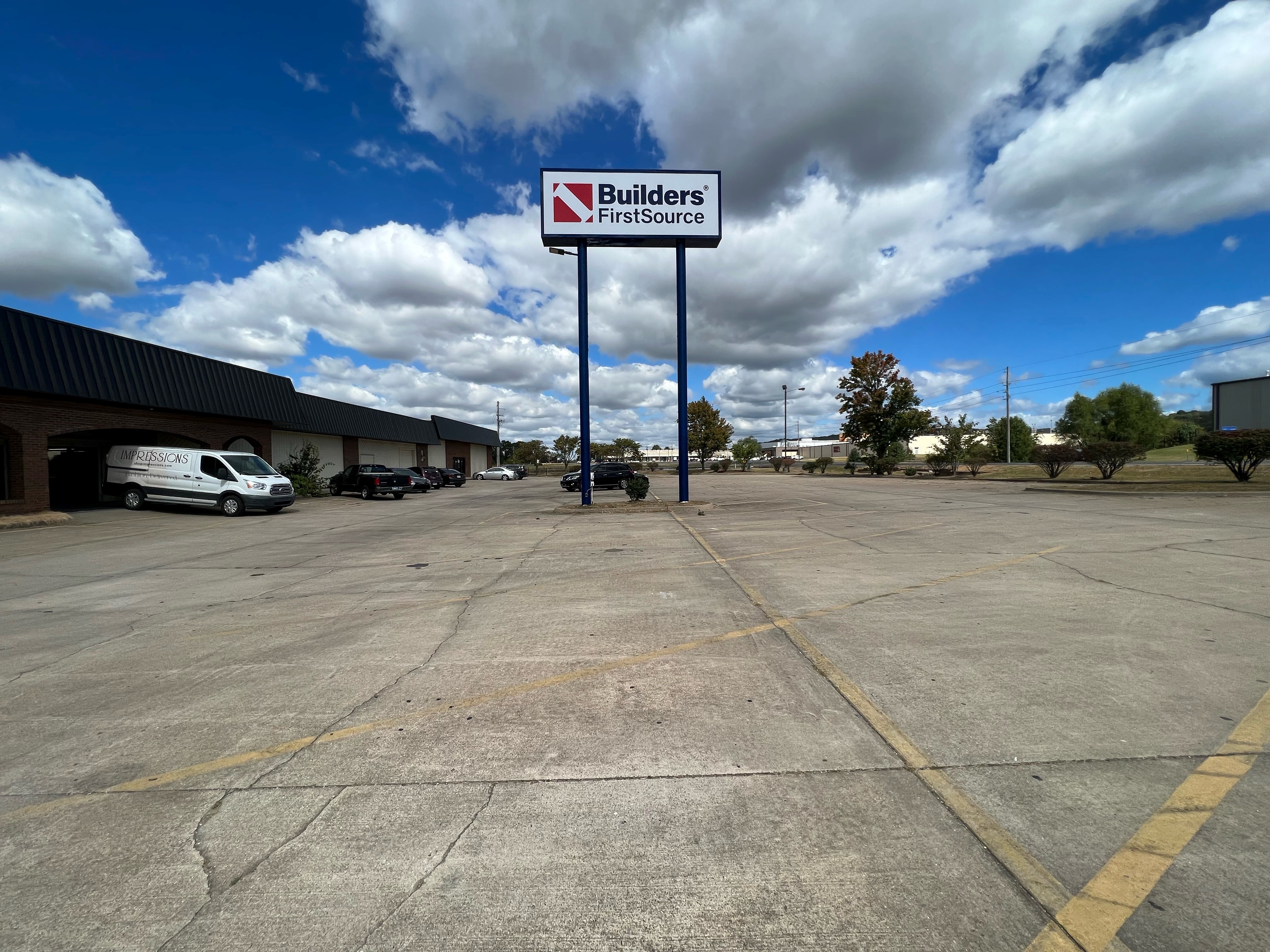 A large parking lot in front of a Builders FirstSource building. The prominent company sign is displayed on a tall pole in the center of the image, with the building on the left side and some parked vehicles, including a white service van. The parking lot is mostly empty, and the sky is filled with puffy clouds against a bright blue background. Sparse trees and bushes line the parking lot.