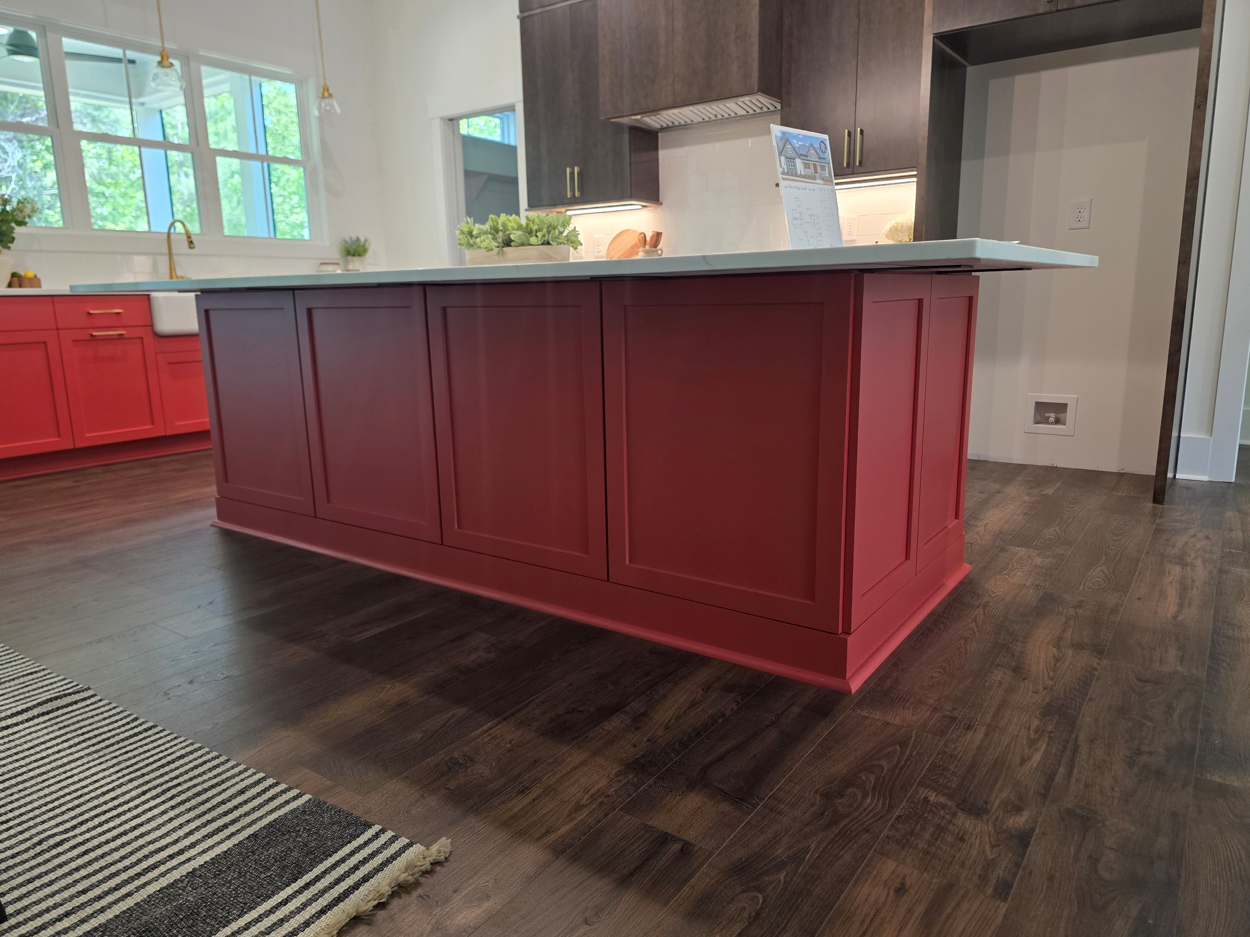 Wide view of a finished kitchen showcasing a large red island with paneled cabinetry, white quartz countertop, dark upper cabinets, stainless steel appliances, and wood flooring.