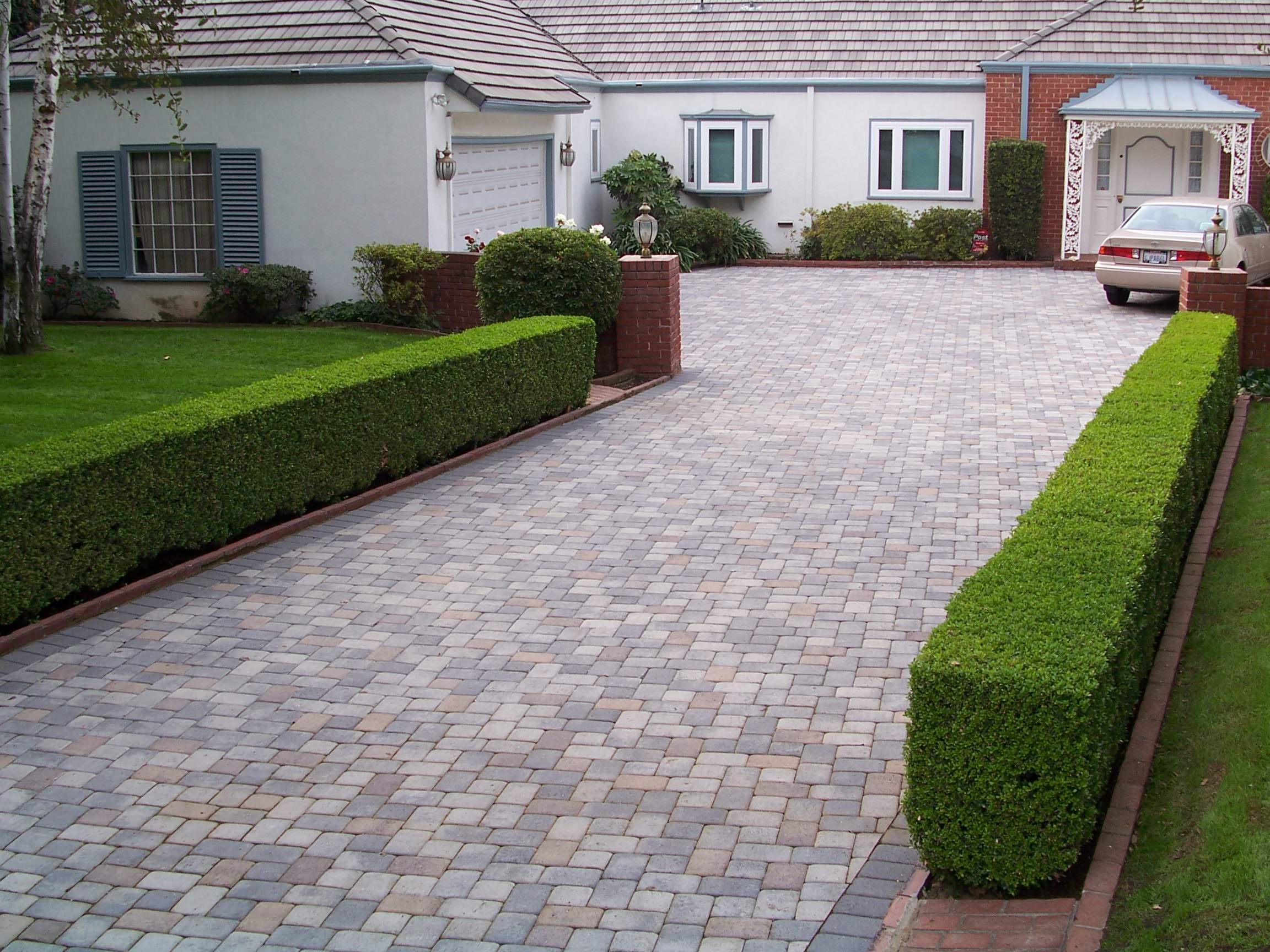 Neatly paved stone driveway bordered by clipped hedges leads to a tidy gray house with garage, parked car, and manicured lawn.