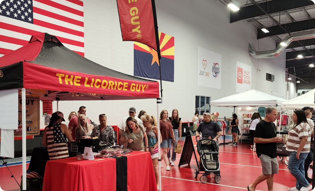 The Licorice Guy booth set up at an indoor fair and market event, featuring the signature red and black branded tent, feather banner flags, and product displays. Customers browse and sample old fashioned licorice at the event.