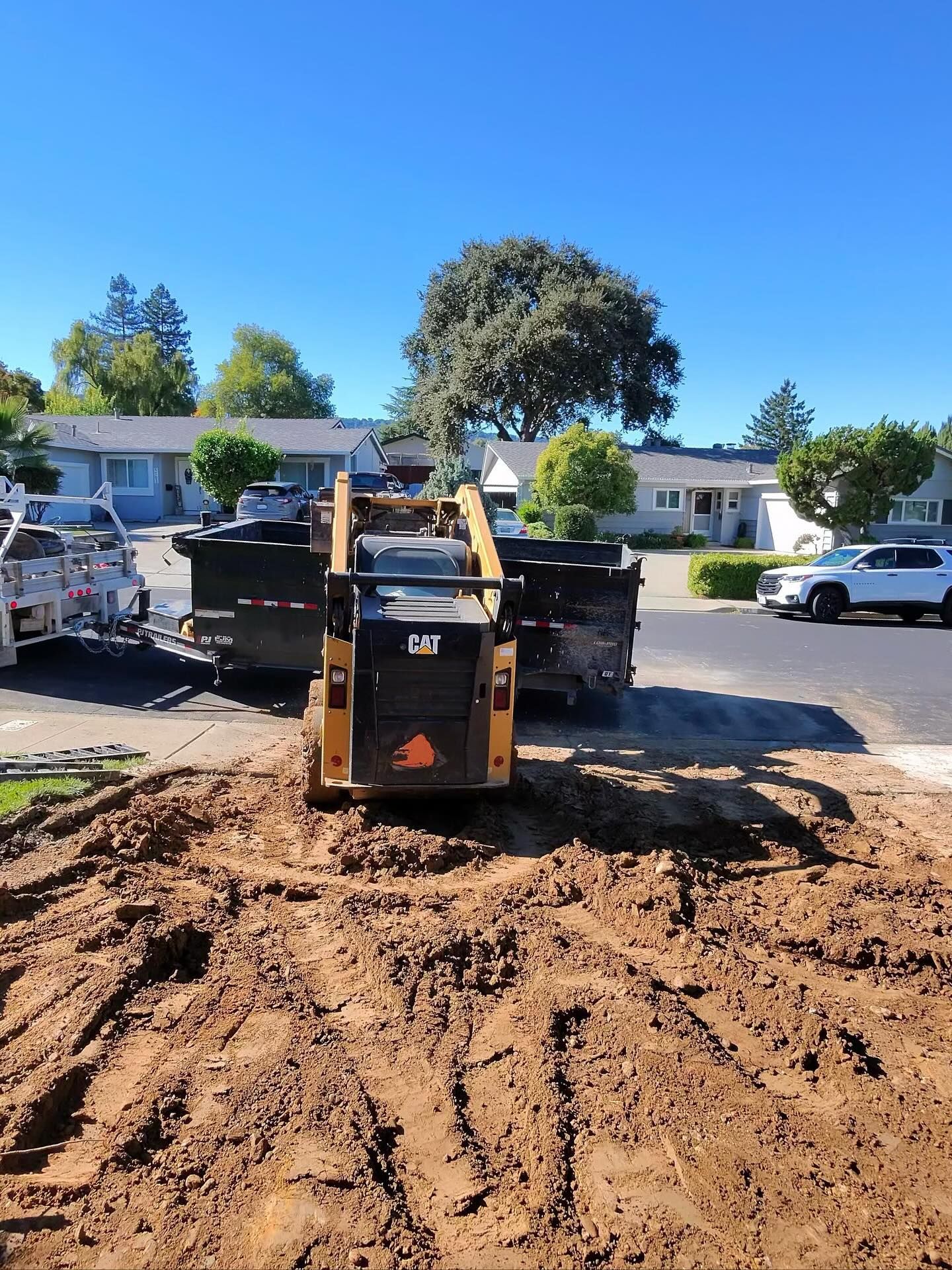 Skid steer loader working on a residential excavation site, moving soil and debris toward a dump trailer, showcasing grading, site preparation, and heavy equipment operation in a suburban neighborhood under clear skies.