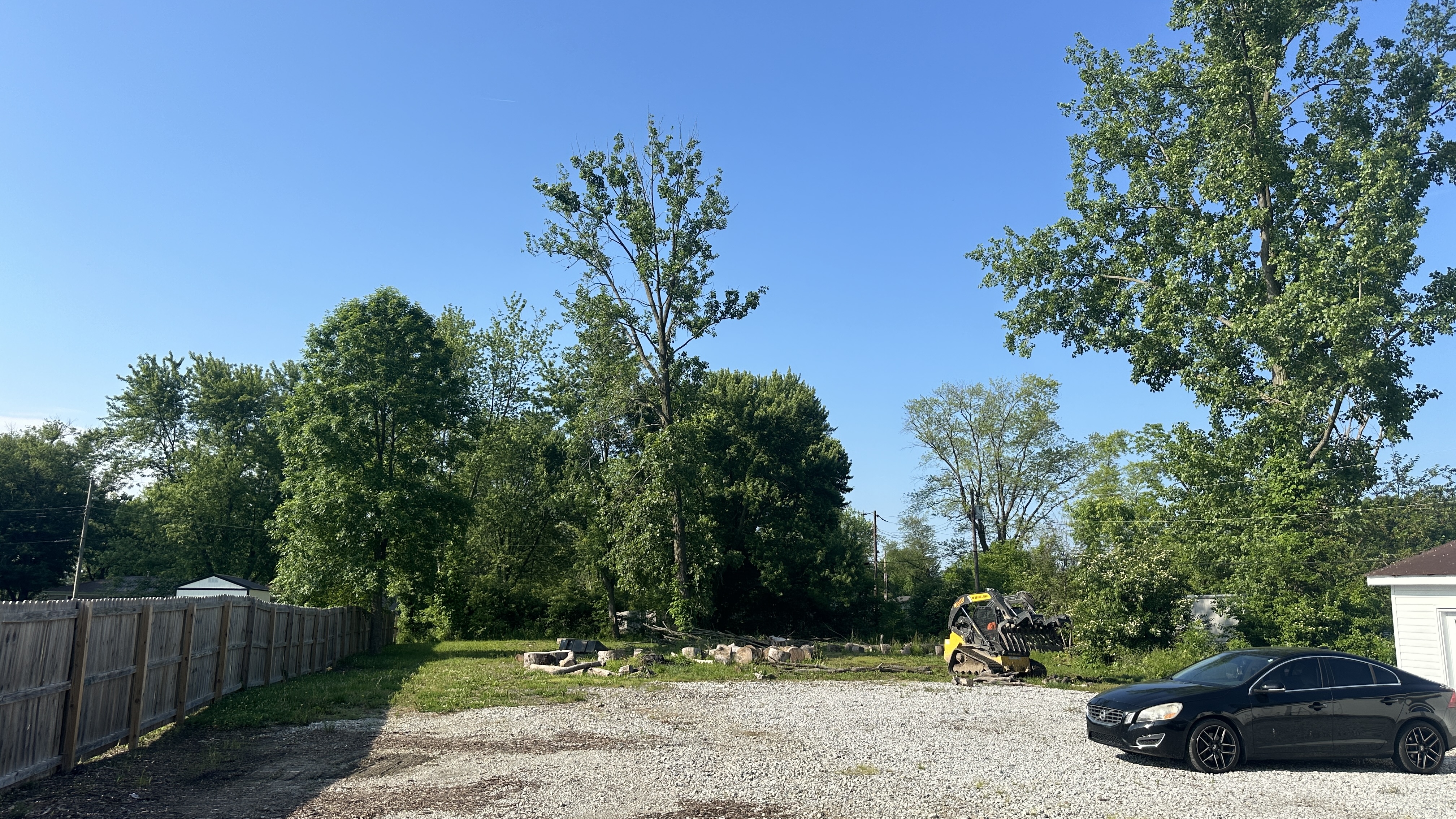 A black Volvo sedan is parked on a gravel lot next to a small construction vehicle. Behind them, a wooden fence encloses a grassy area with felled trees and stumps. Lush green trees and a bright blue sky fill the background.
