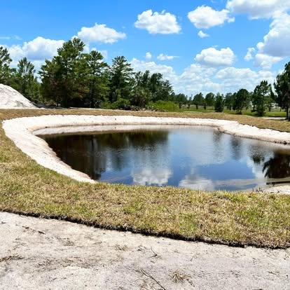 A completed retention pond with dark water, a clean white sand shoreline, and freshly laid green sod along the edge.