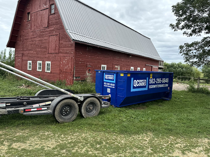 A blue QC Dumpster Dudes dumpster placed next to a red barn in a rural setting, perfect for agricultural or farm cleanups.