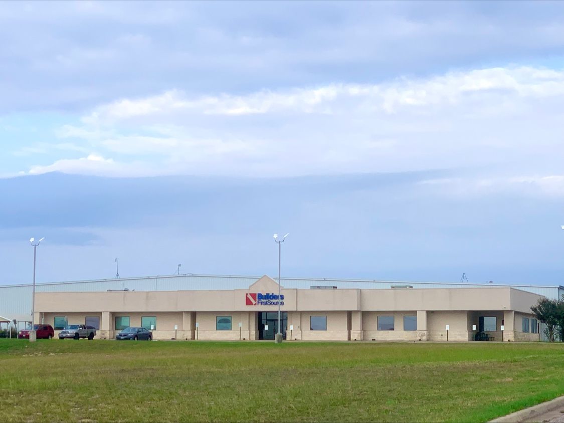 An exterior view of a Builders FirstSource building situated in a large open field. The structure is a single-story industrial-style facility with a beige facade and multiple windows. The company logo is prominently displayed above the entrance, and a few vehicles are parked near the building. The sky is overcast, adding a soft, diffused lighting to the scene.