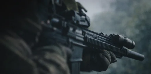 A close-up shot of a soldier's hand holding a rifle with a scope attached, set in an outdoor environment with a blurred background of trees and fog.