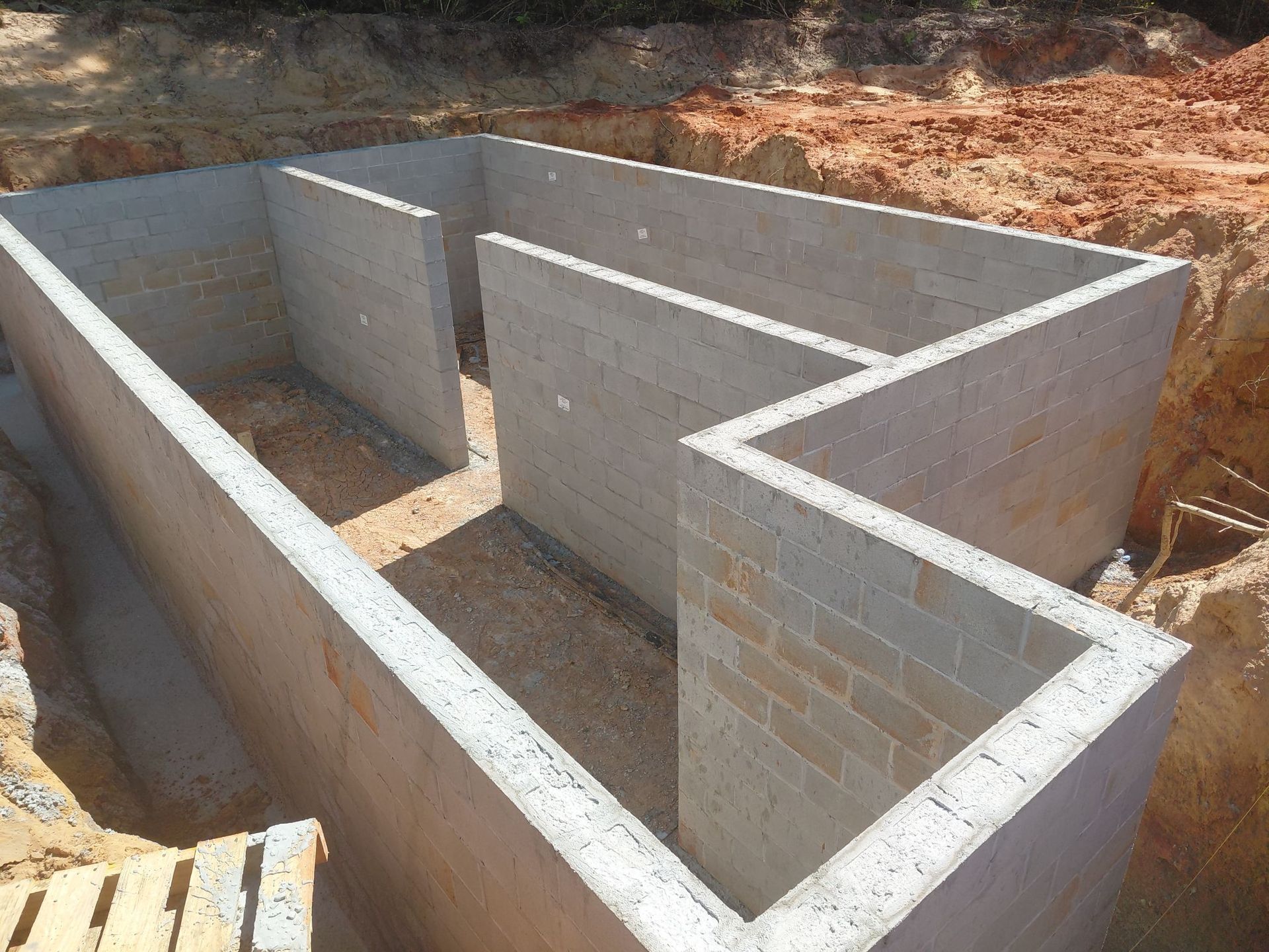 Concrete block foundation walls under construction forming basement layout, with multiple interior partitions, excavated soil surrounding structure, and unfinished floor base inside rectangular perimeter of newly built foundation.