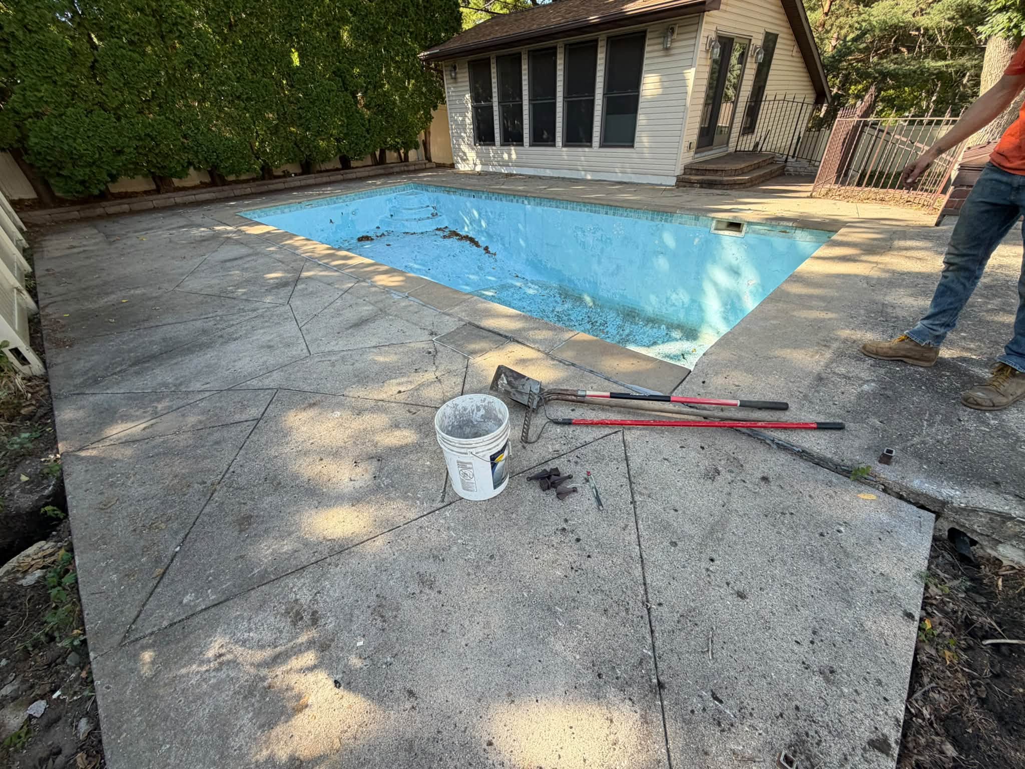 A wide shot of an drained, empty inground swimming pool surrounded by a decorative, geometrically scored concrete deck. Maintenance tools, including a white five-gallon bucket, shovels, and squeegees, rest on the concrete in the foreground alongside a worker standing nearby. In the background sits a pool house with large windows and a privacy fence line.