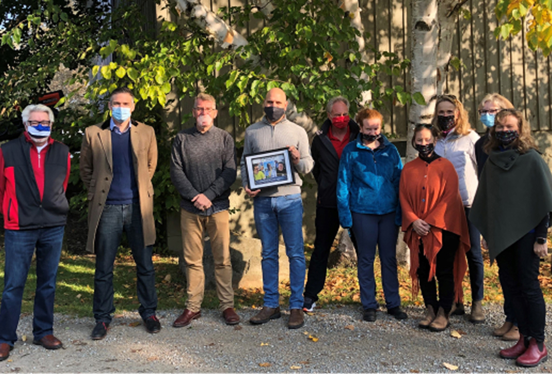 A group of eleven people stands outdoors, all wearing masks. One person holds a framed picture. They are surrounded by green trees, conveying an autumn setting.
