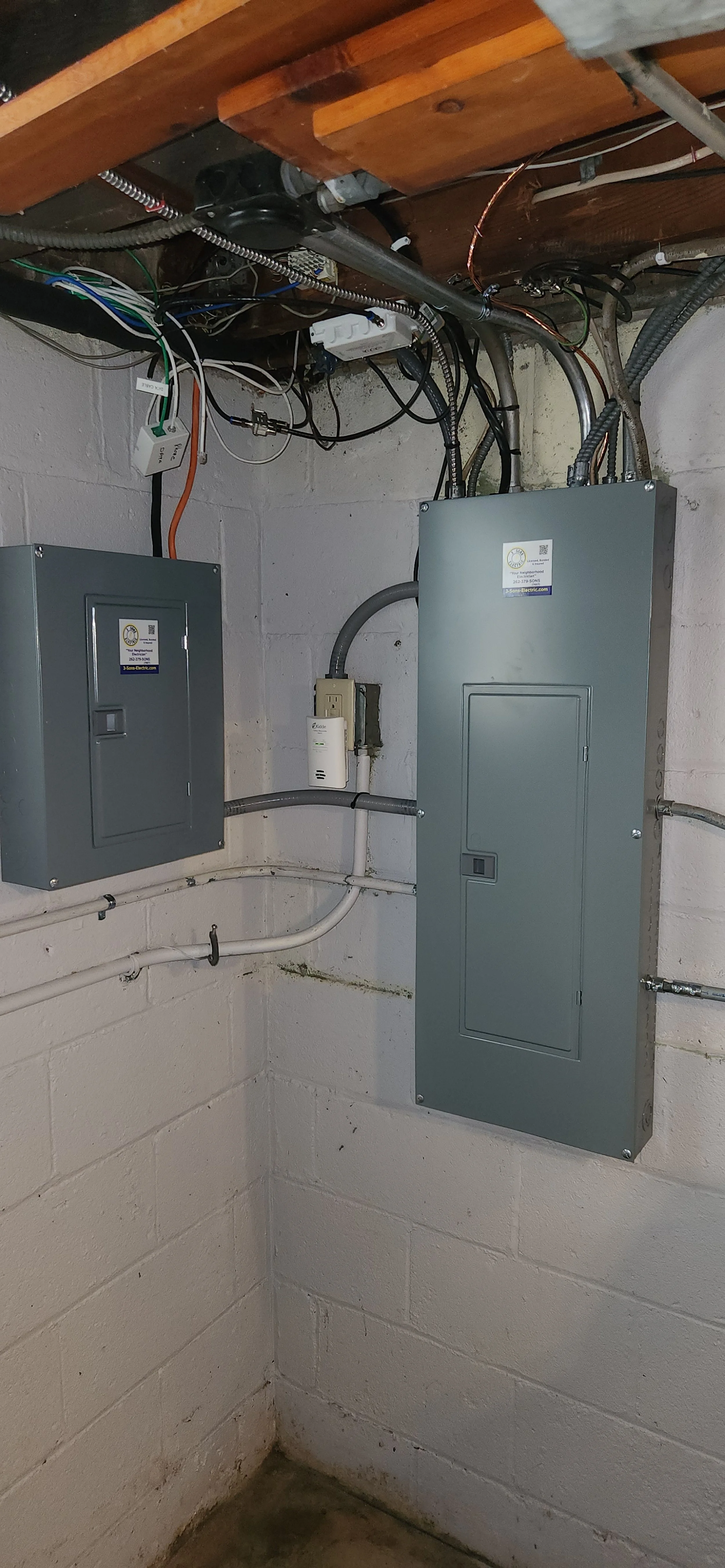 Two gray electrical panels are mounted on a white brick wall in a basement. The panel on the left is smaller and mounted higher than the larger panel on the right. Both panels have small doors with latches. The larger panel on the right has a sticker with a logo and text on its door. Wires and conduits run along the ceiling and the walls, connecting to the panels and other electrical components. The ceiling is made of wooden beams and has various electrical wiring and pipes running across it. The floor is a dark concrete surface.