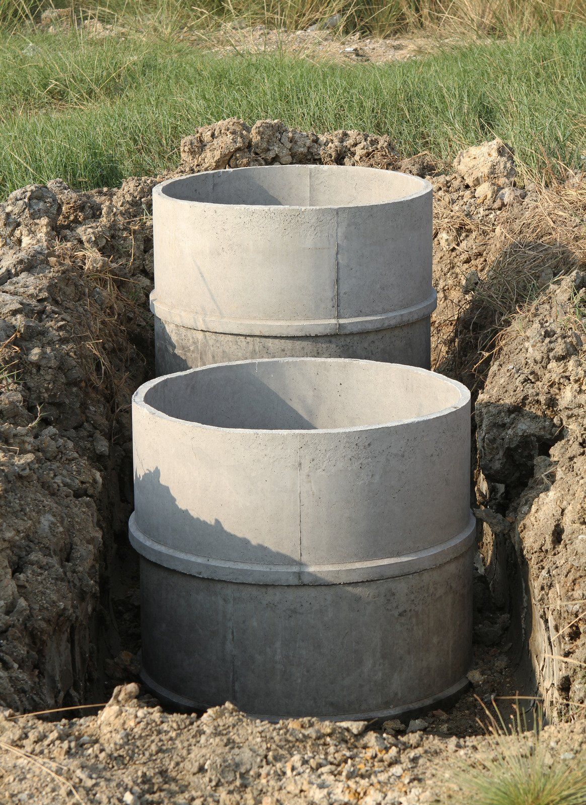 Two large, circular precast concrete cylinders are installed vertically within a narrow earthen trench. These heavy-duty riser sections are stacked and aligned to form a subterranean drainage or utility access point.
