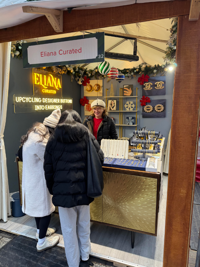 Discover Eliana Curated&rsquo;s festive jewelry booth, featuring earrings crafted from upcycled designer buttons, necklaces and bracelets. Shoppers explore unique accessories surrounded by holiday greenery.