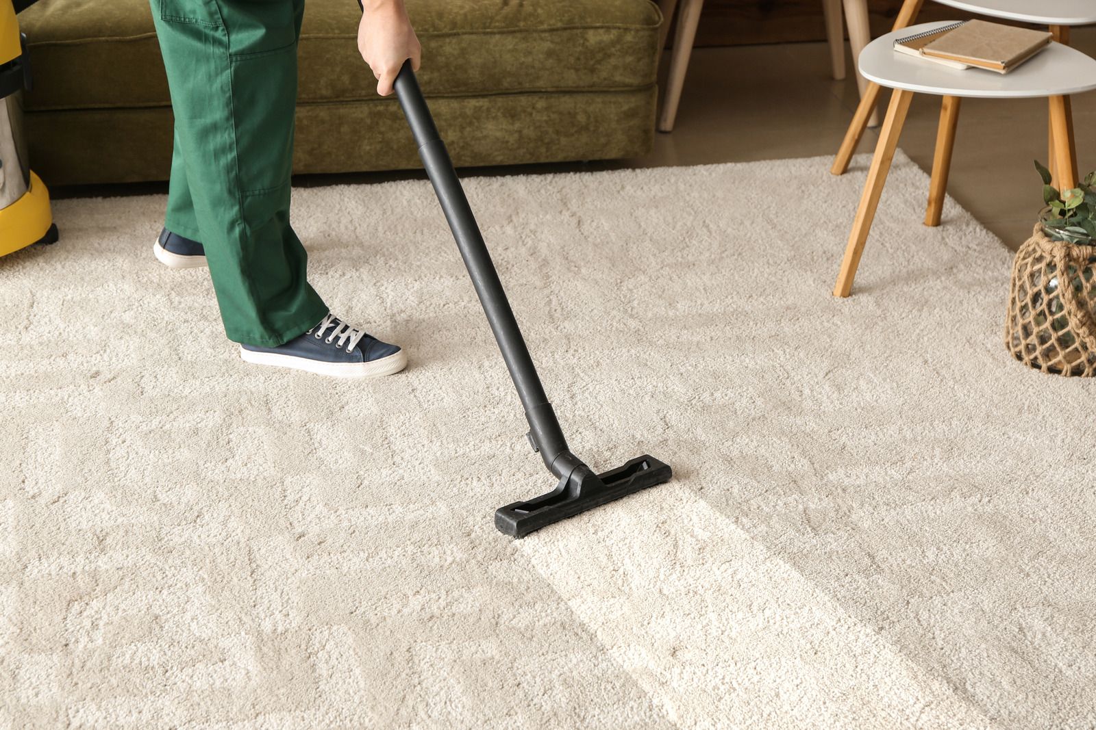 Person vacuuming light-colored carpet in living room, creating visible clean lines, with sofa and side table nearby, highlighting routine carpet cleaning, maintenance, and tidy residential interior environment.