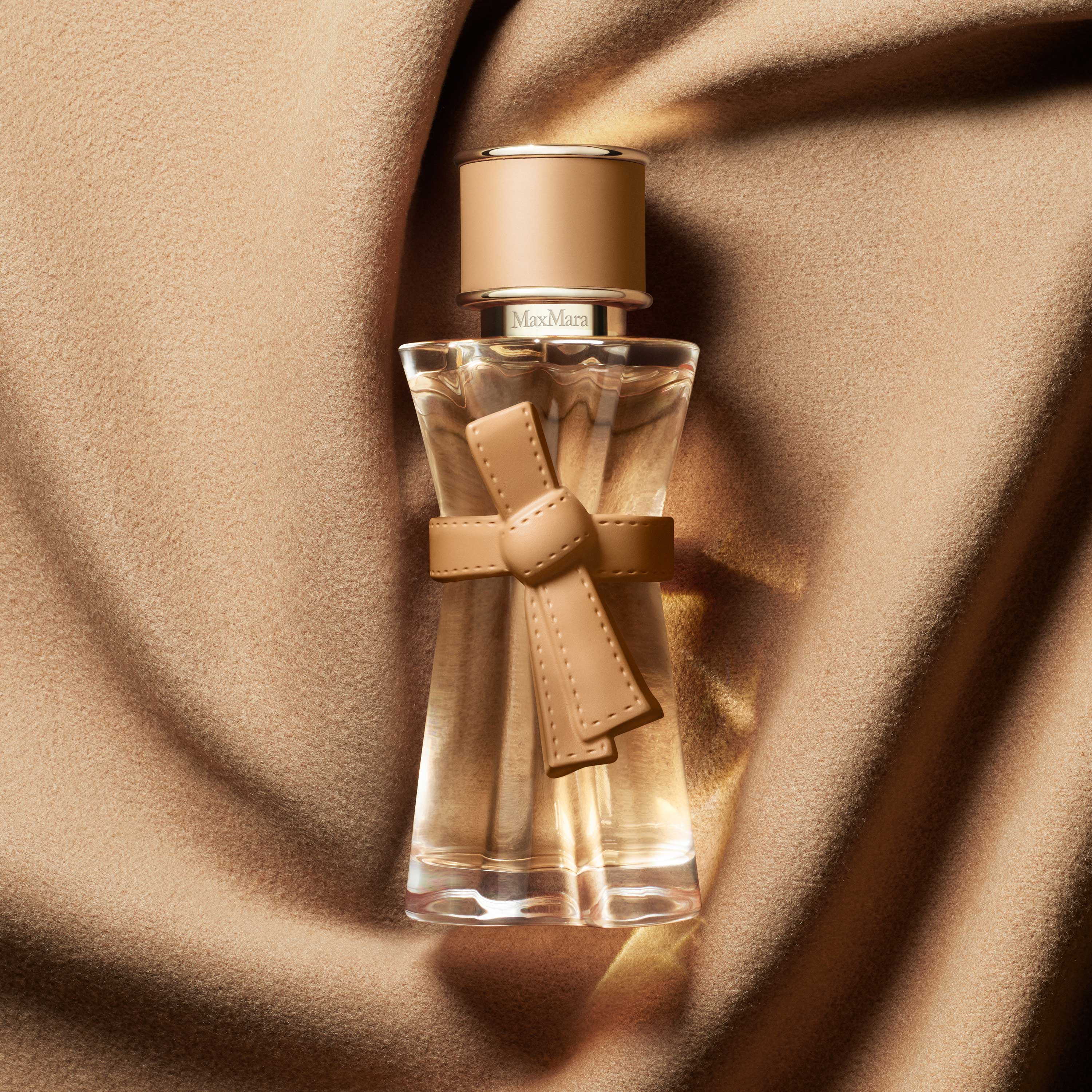 Un frasco de perfume Max Mara con un lazo color camello, colocado sobre una lujosa tela de lana drapeada.