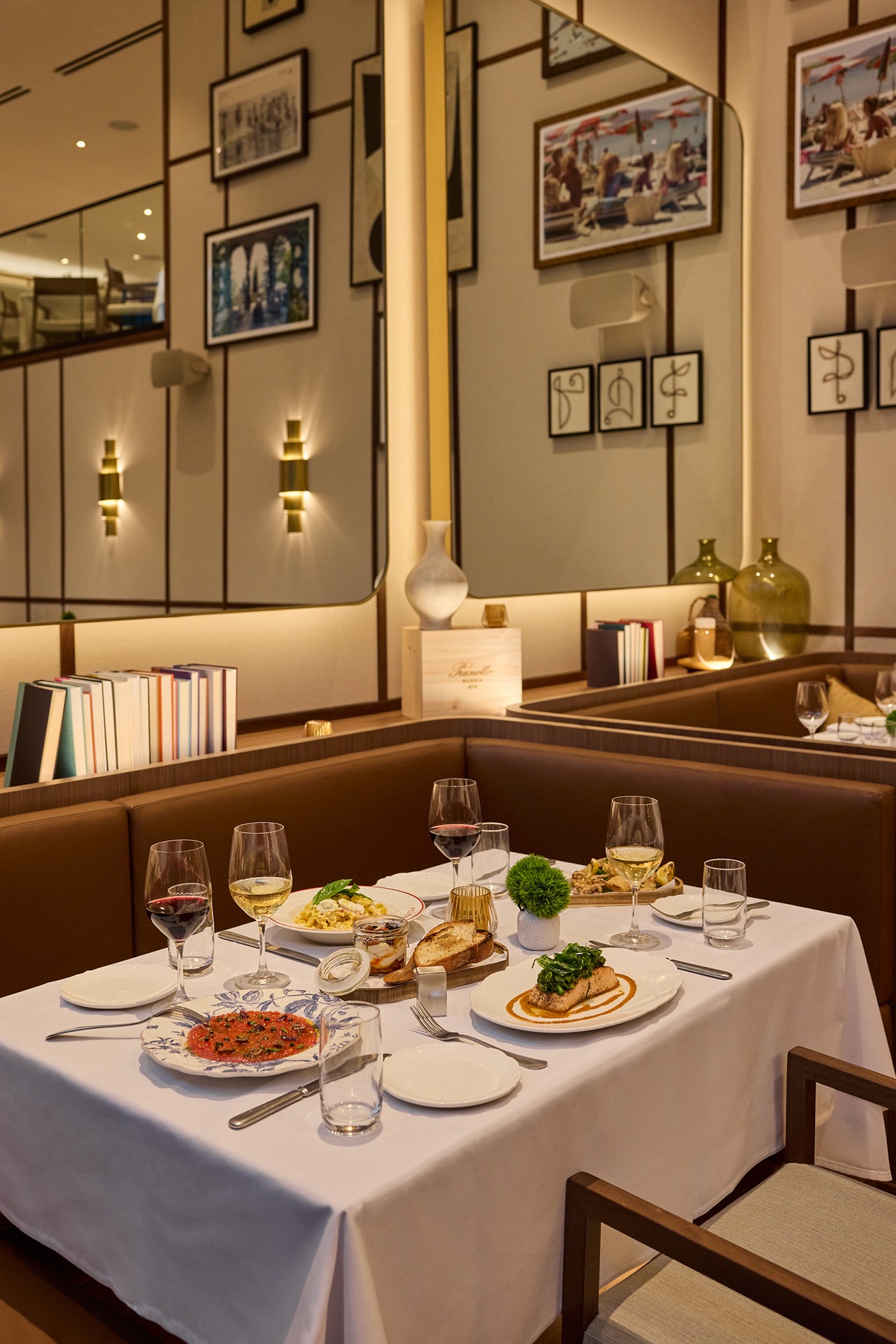 A white-clothed table set with Italian entrees and wine glasses. The background features large mirrors and framed photography, creating a warm, upscale atmosphere.