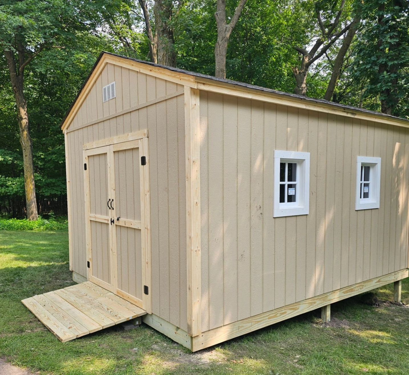 A finished, long storage shed with light tan vertical siding and a gable roof, set in a wooded backyard. It features double wooden doors with a ramp, and two white-trimmed windows along the side.