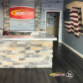 Signstore reception area featuring a stone counter and a large Signstore logo on the wall behind it. The area has a wood-like floor, and a decorative American flag is displayed on the right wall. The "26" anniversary badge is visible in the corner.