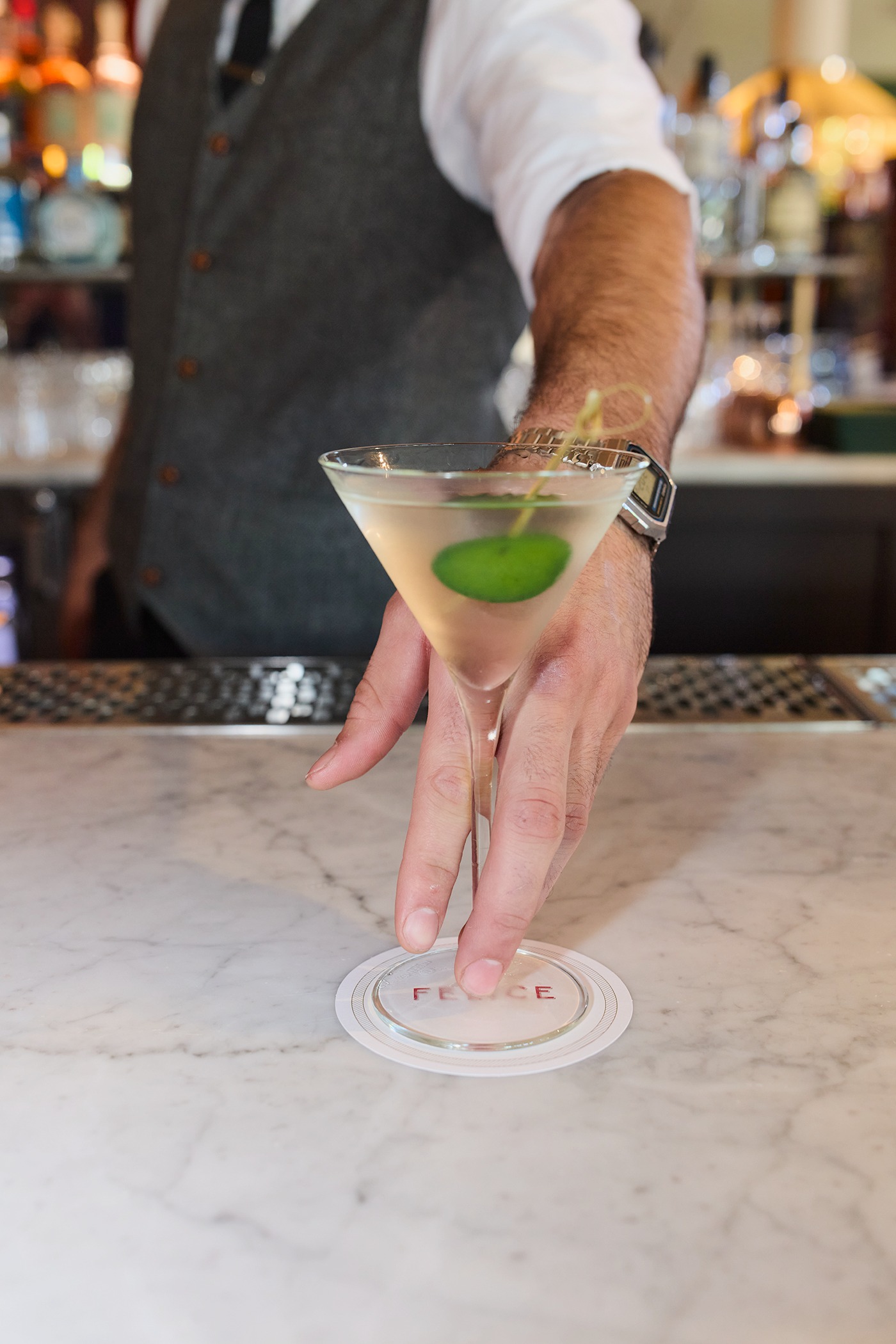 A close-up of a bartender&rsquo;s hand placing a clear martini garnished with a large green olive onto a coaster. The bartender wears a grey wool vest and a white shirt against a backdrop of liquor bottles.