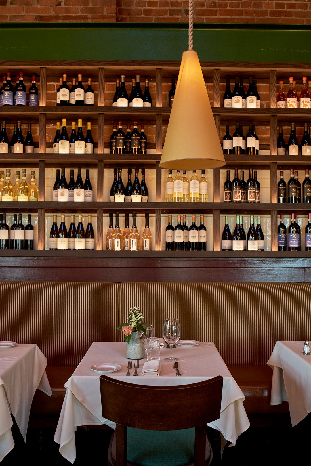 A table for two with white linens and a small floral centerpiece is centered in front of a massive wooden wine library. A large conical pendant lamp hangs low over the table, creating an intimate atmosphere.