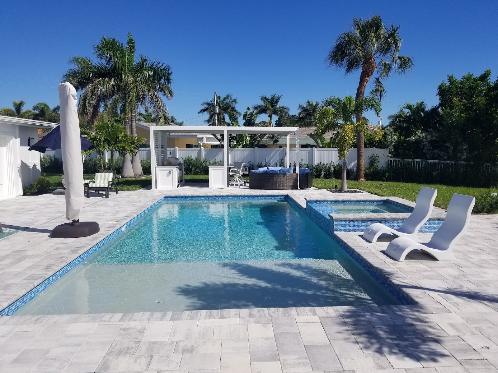 Modern rectangular backyard swimming pool with clear blue water, light stone pavers, lounge chairs, and palm trees.
