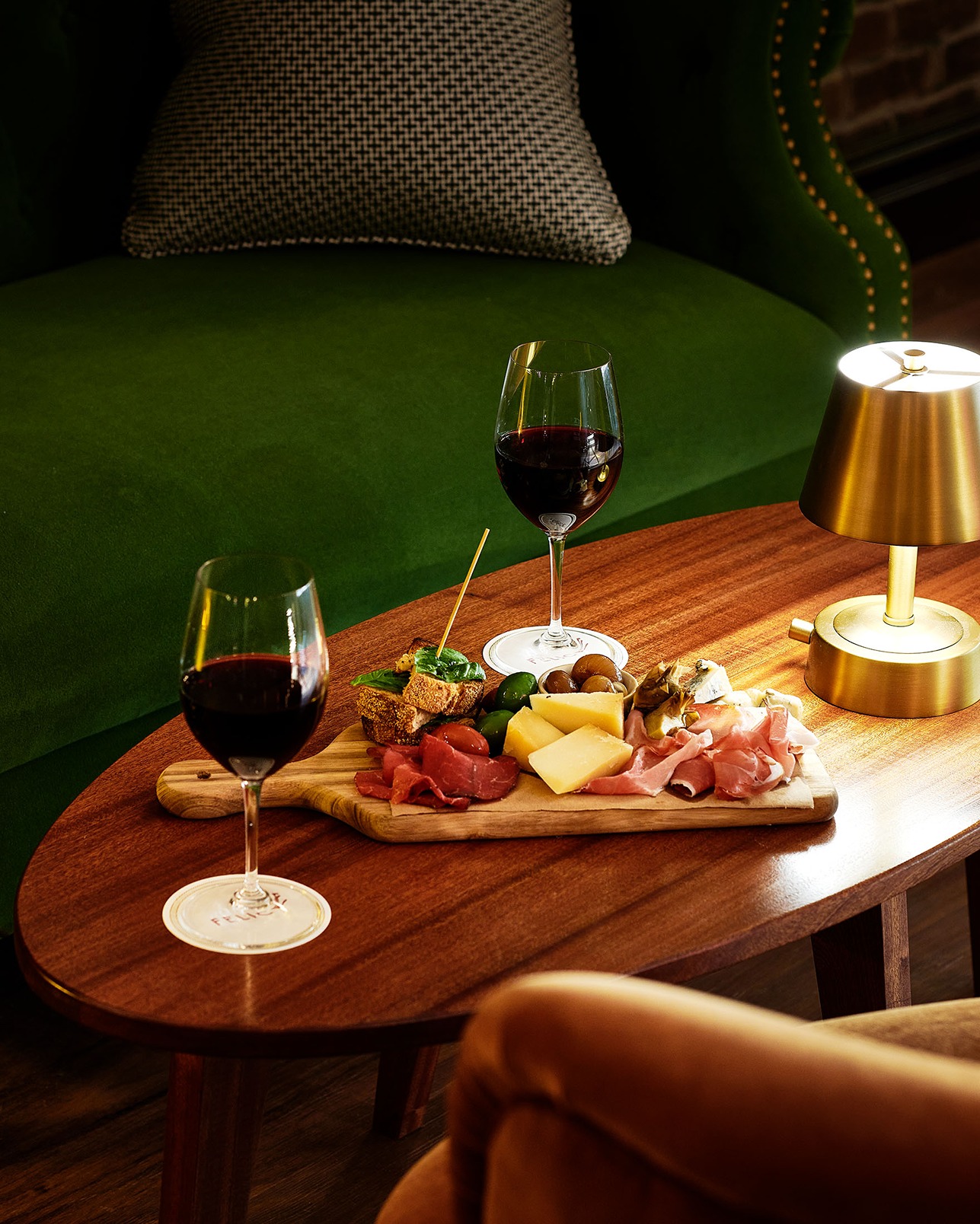 A charcuterie board with cheeses, olives, and meats sits on an oval wooden table next to two glasses of red wine and a modern gold lamp, positioned in front of a green velvet sofa.