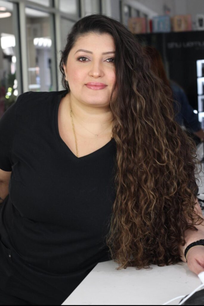 A close-up, medium shot of a woman with long, curly brown hair and makeup, wearing a black v-neck t-shirt, smiling. She is leaning on a white surface and looking at the camera. The background is blurred, showing a window with reflections and some shelves with items on them.