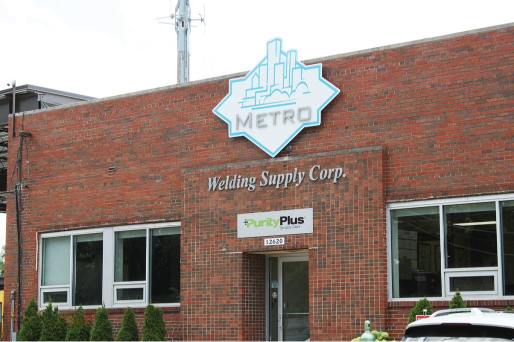 This image shows the Metro Welding Supply Corporation main office building, a brick commercial structure with signage indicating its business operations.

Key Features of the Image:

Large "Metro Welding Supply Corp." Signage – The company name is prominently displayed on the front brick facade in metallic silver letters.
Metro Logo – Above the company name is the Metro Welding Supply logo, featuring a stylized city skyline inside a blue diamond shape, symbolizing its connection to the Detroit, Michigan area.

PurityPlus Specialty Gases Sign – A PurityPlus Specialty Gases logo is mounted near the entrance, indicating that Metro Welding Supply is an authorized distributor of high-purity specialty gases.

Building Details – The exterior is made of red brick, with large front windows and a glass entrance door with the address 12620 displayed above it.
Surrounding Area – A row of neatly trimmed green shrubs is planted in front of the building, and a vehicle with gas cylinders is partially visible in the lower right corner, indicating active business operations.

This professional and industrial-looking facility serves as the headquarters for Metro Welding Supply Corp., a trusted supplier of specialty gases, welding equipment, and industrial solutions in Detroit, MI and surrounding areas.