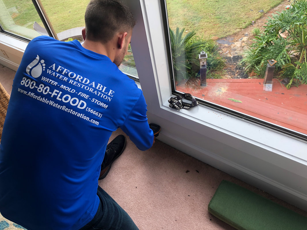A man wearing a blue shirt with the logo "AFFORDABLE WATER RESTORATION" is using a moisture meter near a window. The shirt also displays the phone number "800-80-FLOOD" and the website "www.AffordableWaterRestoration.com". The scene appears to be inside a home, possibly in a living room or bedroom, with a view of an outdoor deck and greenery through the window. A pair of work gloves is visible on the windowsill.