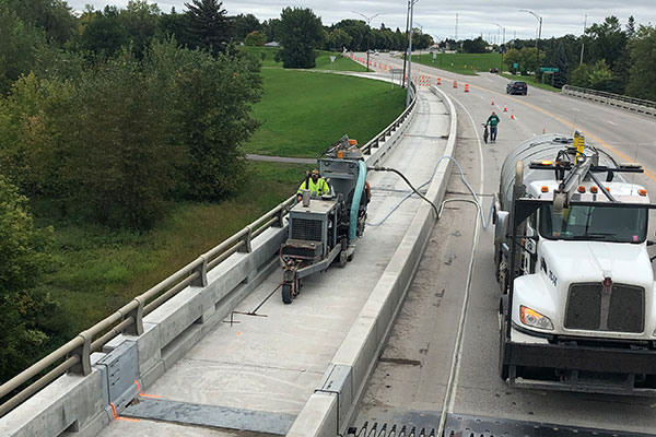 An elevated, wide-angle aerial perspective photograph taken from a pedestrian walkway looking down onto a bridge deck under construction. Centered on the operational deck is a DSI specialty bridge deck grinding machine actively operating in a closed lane, with a worker in high-visibility gear near the controls. A large white Kenworth DSI support tanker truck is parked adjacent to it, supplying water or managing slurry. The bridge spans a wooded area, and active traffic flows on the far right lane.