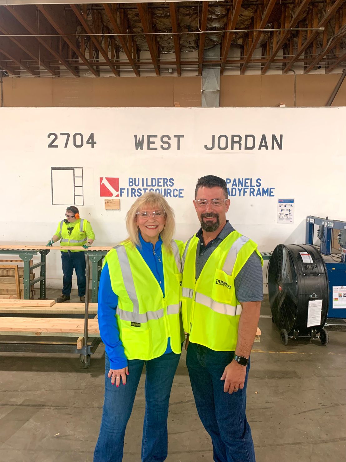 Two employees wearing high-visibility safety vests standing inside the Builders FirstSource READY-FRAME facility in West Jordan, UT. Behind them, a worker operating a machine near stacks of lumber under a sign reading '2704 West Jordan' and company branding.