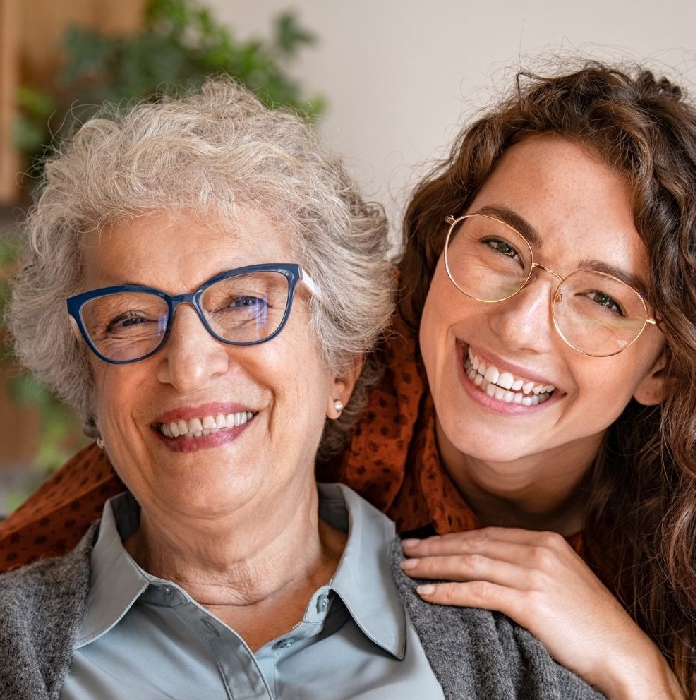 A warm indoor portrait of two women facing the camera. In front, an older woman with short silver curls, navy cat-eye glasses, a pale blue button-down, and a gray cardigan smiles broadly. Behind her, a younger woman with long wavy brown hair, gold wire-frame glasses, and a rust-patterned scarf leans in with her hand resting on the older woman's shoulder. Soft green houseplant is blurred in the background.