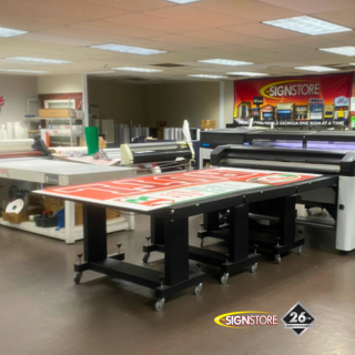 Signstore vinyl production department featuring large printing tables, printers, and a workstation with various signage materials. The area is well-organized and designed for high-volume production. The "26" anniversary badge is visible.