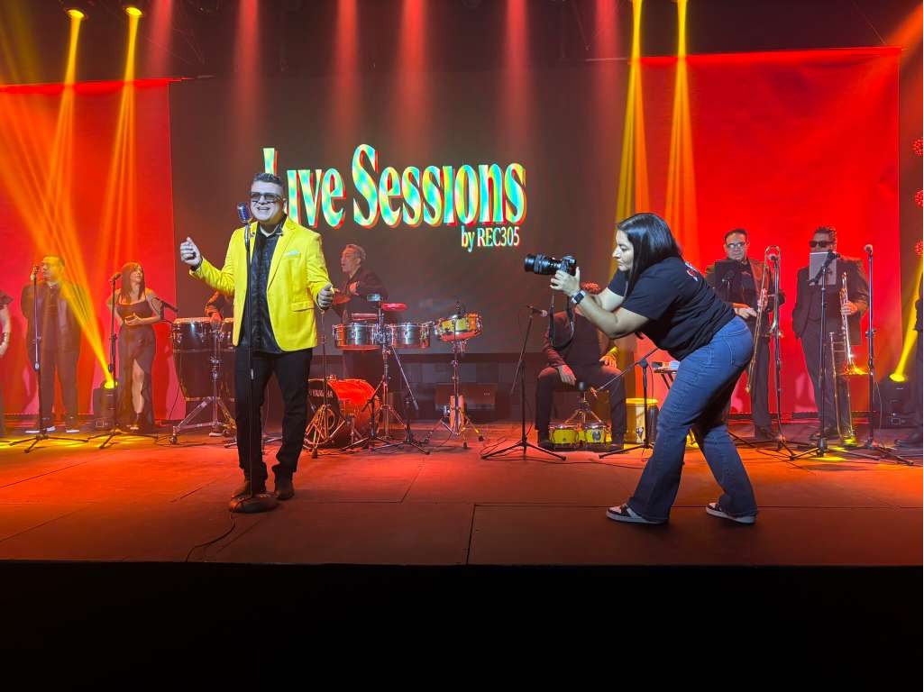 En esta toma cercana te mostramos la energ&iacute;a sobre nuestra tarima. Se observa a un cantante con saco amarillo interpretando un tema junto a su banda de vientos, bajo una c&aacute;lida iluminaci&oacute;n en tonos amarillos y rojos. En el lado derecho, una fot&oacute;grafa de nuestro equipo se agacha para capturar la toma perfecta, mientras la pantalla central proyecta el logo del evento con total nitidez. Somos el sal&oacute;n de fiesta con todo incluido que est&aacute;s buscando. Escr&iacute;benos para enviarte una cotizaci&oacute;n de sal&oacute;n de eventos.