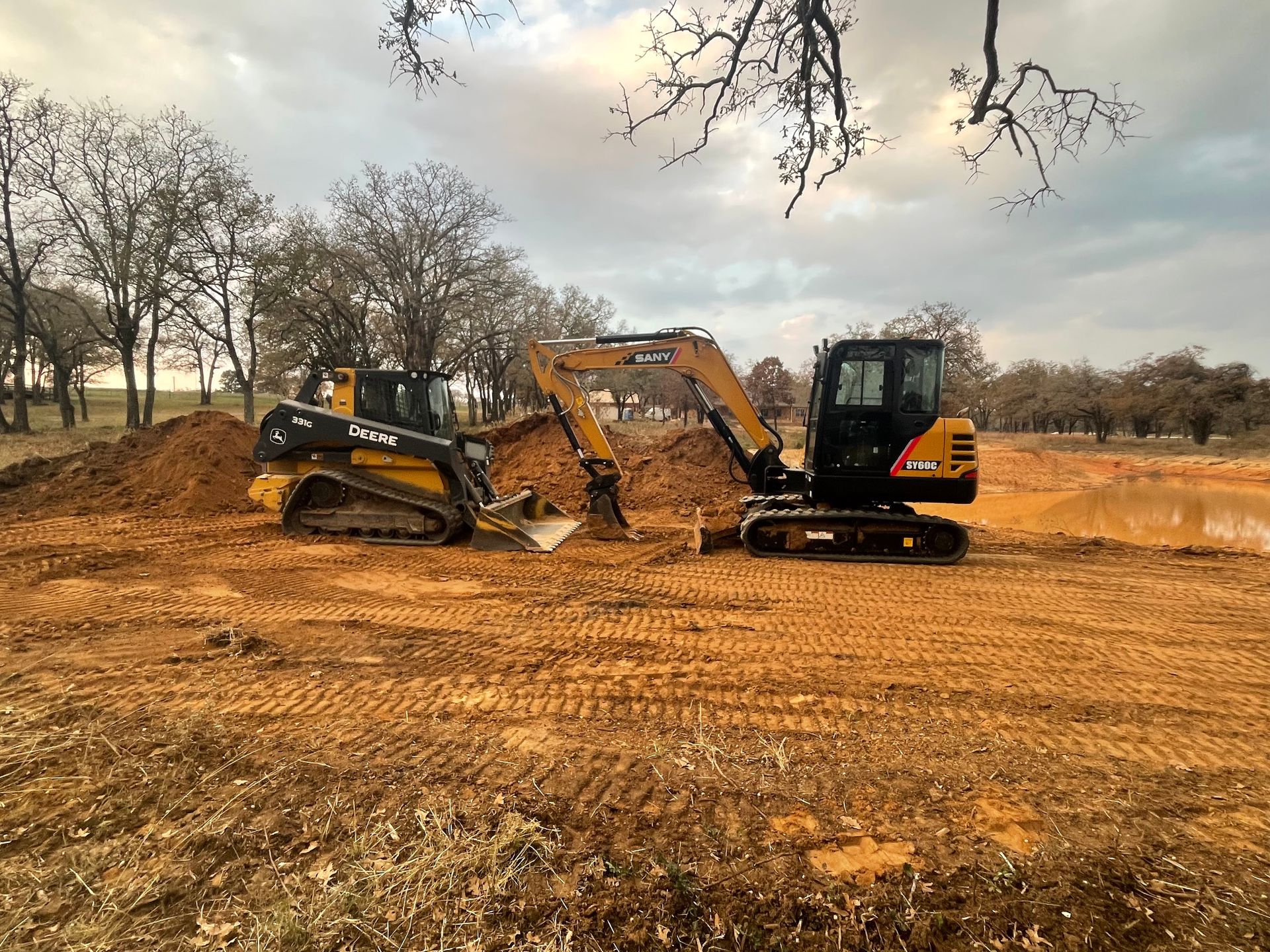 Excavator and skid steer working near pond excavation site, moving soil and shaping terrain, showcasing pond construction, excavation, and grading services for land development and water feature installation projects.