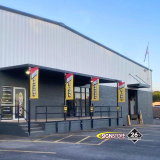 Exterior of Signstore building featuring modern white and gray design. Vertical banners read “SIGNSTORE” and “SIGN CENTER.” The entrance has a ramp and stairs. Signstore logo and "26" anniversary badge visible.