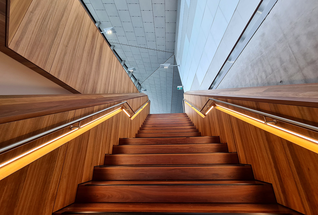Wide wooden staircase ascending between modern, angular walls. Warm lighting under handrails creates a welcoming, futuristic ambiance.