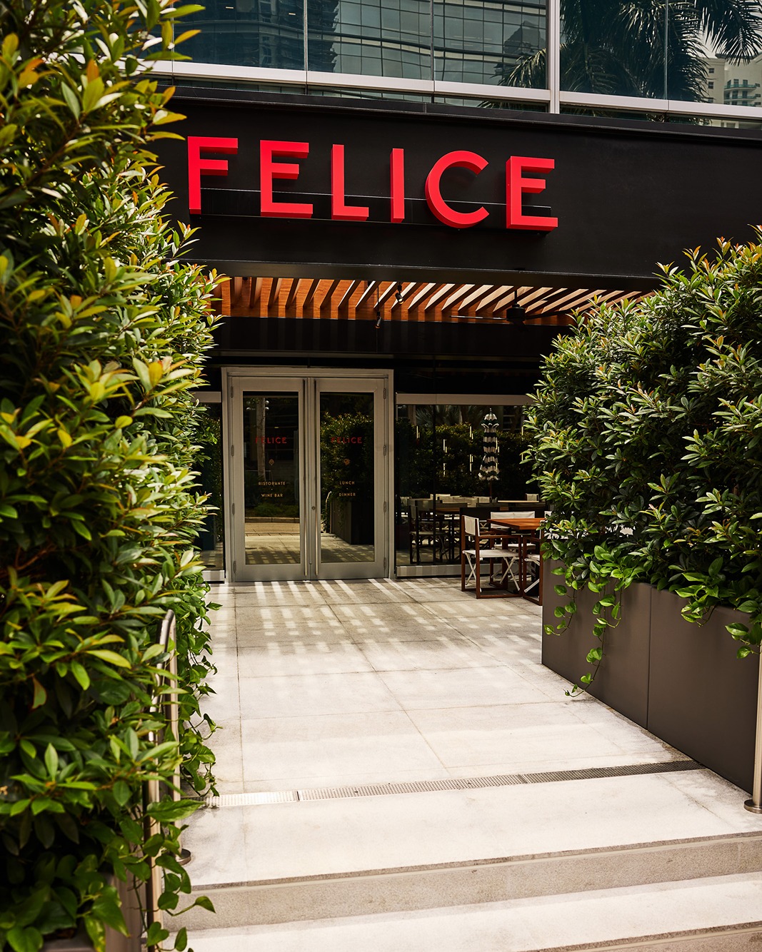A sleek black storefront with a vibrant red "FELICE" sign. Modern glass doors lead inside, framed by lush green manicured hedges and a sun-drenched stone walkway.